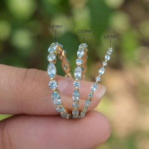 May include: Three gold rings with light blue gemstones of varying sizes. The rings are held by a person's fingers. The text on the image indicates the gemstone sizes are 2.5 mm, 2.0 mm, and 1.5 mm.