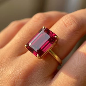 May include: A gold ring with a large, rectangular, deep red gemstone. The gemstone is set in a gold band with four prongs. The ring is displayed on a finger.