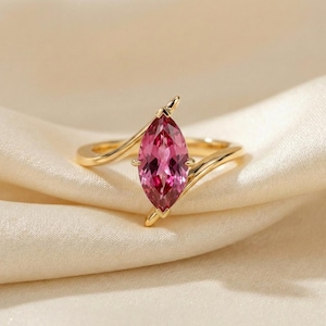 Op de afbeelding: Een gouden ring met een roze edelsteen in marquise-vorm. De edelsteen is het middelpunt, gezet in een delicate, golvende gouden band. De ring wordt gepresenteerd op een zachte, crèmekleurige stof, wat het elegante ontwerp benadrukt.