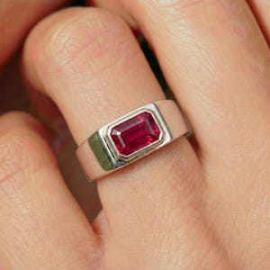 May include: A silver ring with a rectangular, deep red gemstone. The gemstone is set in a bezel setting, and the ring band is wide and polished. The ring is on a finger.