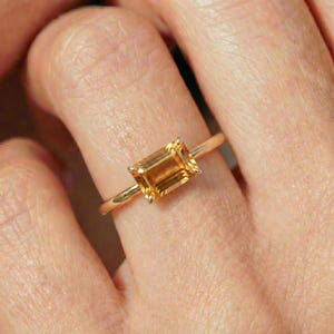 May include: A gold ring with a rectangular, amber-coloured gemstone. The ring's band is thin and gold, with the gemstone set in four prongs. The gemstone has a warm, golden hue, and the ring is shown on a finger.