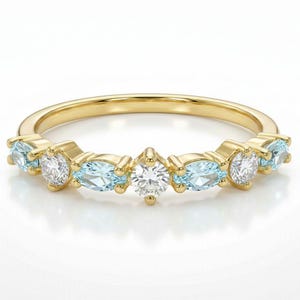 May include: A gold band ring featuring a series of alternating gemstones. The ring has oval-cut light blue gemstones and round, clear gemstones. The ring is set on a white background.