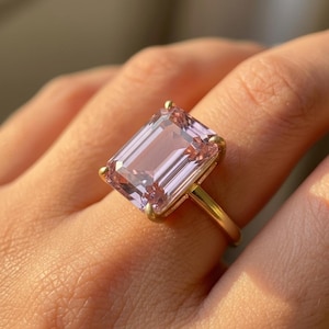 Può includere: Un anello in oro con una grande pietra preziosa rettangolare color rosa chiaro, taglio smeraldo, fissata con quattro griffe. La pietra ha una superficie sfaccettata e limpida che riflette la luce, mentre la fascia è in metallo dorato liscio.