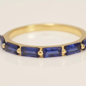 May include: A gold band ring featuring rectangular, deep blue gemstones. The ring has a simple design with the gemstones set in a row, separated by small gold beads. The ring is a piece of jewelry.