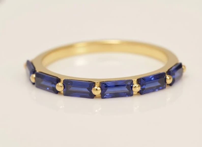 May include: A gold band ring featuring rectangular, deep blue gemstones. The ring has a simple design with the gemstones set in a row, separated by small gold beads. The ring is a piece of jewelry.
