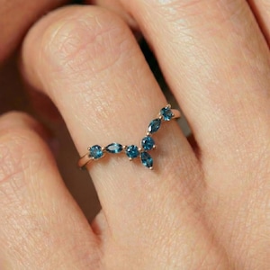 Puede incluir: Un anillo dorado con un diseño curvo, adornado con múltiples piedras preciosas azules. Las piedras son una mezcla de tallas redondas y marquesa, creando una apariencia delicada y elegante. El anillo se muestra en un dedo.