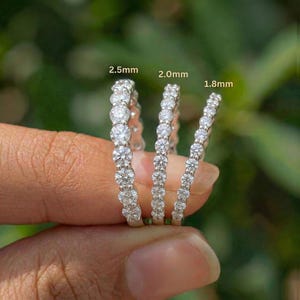 May include: Three silver rings with clear, round gemstones are displayed. The rings are labeled with sizes: 2.5mm, 2.0mm, and 1.8mm. The rings are held against a blurred green background, showcasing the sparkle of the gemstones.