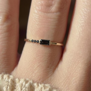 Könnte beinhalten: Ein zarter Goldring mit einem rechteckigen schwarzen Edelstein und drei kleineren schwarzen Edelsteinen. Der Ring hat ein dünnes Band und ein minimalistisches Design. Die Edelsteine sind in einer linearen Anordnung angeordnet.