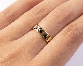 May include: Gold band ring with a mountain range design. The ring is worn on a finger.