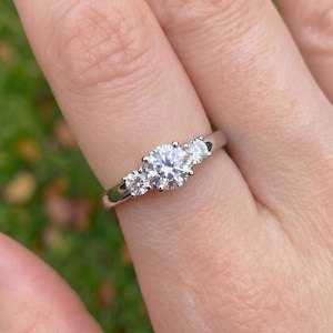 May include: A three-stone ring featuring a large round brilliant-cut center diamond flanked by two smaller round diamonds. The stones are set in a polished silver-toned metal band that encircles the finger, showcasing a classic design.