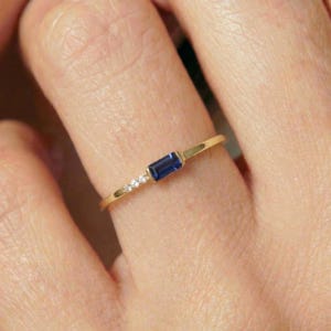 May include: A delicate gold ring featuring a rectangular, deep blue gemstone and three smaller, clear gemstones. The ring has a thin band and is worn on a finger, showcasing its minimalist design and elegant aesthetic.