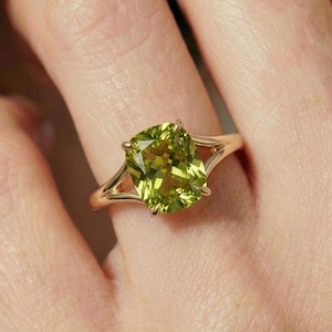 Pode incluir: Um anel de ouro com uma grande pedra preciosa verde clara com corte almofada. A banda do anel divide-se num design de banda dupla em ambos os lados da pedra preciosa. A pedra preciosa tem cerca de 1,9 cm de largura.