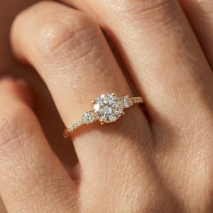 Puede incluir: Un anillo de compromiso de oro con una piedra preciosa grande, redonda y transparente en el centro, flanqueada por piedras más pequeñas. La banda también está adornada con pequeñas piedras preciosas. El anillo está en un dedo.