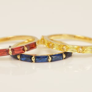 May include: Three gold rings with colorful gemstones. One ring features red gemstones, another has blue gemstones, and the third ring is adorned with yellow gemstones. The rings are set against a white background.