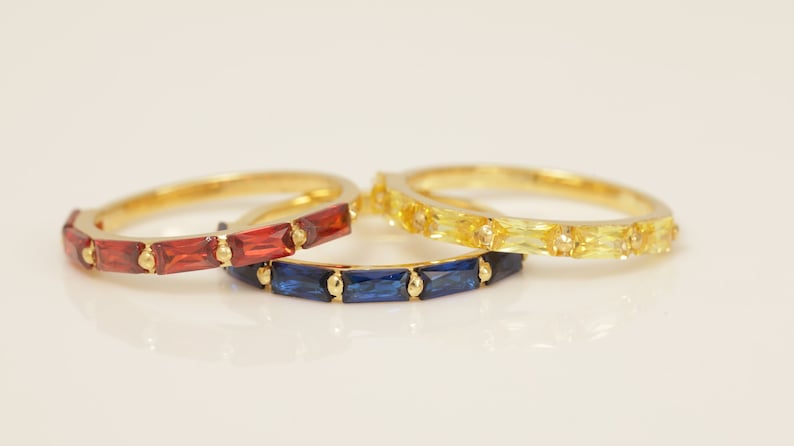 May include: Three gold rings with colorful gemstones. One ring features red gemstones, another has blue gemstones, and the third ring is adorned with yellow gemstones. The rings are set against a white background.