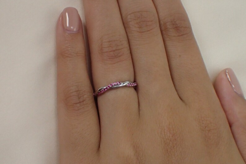 May include: A silver-colored ring with a twisted design, adorned with small, bright pink gemstones. The ring is worn on a finger, showcasing its delicate and intricate details. The gemstones are set closely together, creating a continuous line of color.