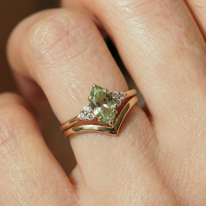May include: A gold ring set featuring a marquise-cut, light green gemstone with small, clear accent stones. The ring is paired with a chevron-shaped band, creating a layered look. The ring is shown on a hand.