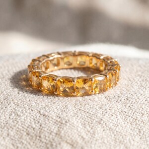 Puede incluir: Un anillo de oro con una banda continua de piedras preciosas amarillas de corte cuadrado. El anillo está colocado sobre una superficie neutra y texturizada. El diseño del anillo es de estilo eterno clásico.