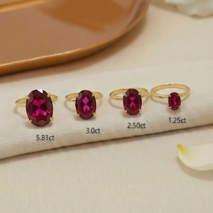 May include: Four gold rings with oval, deep red gemstones of varying sizes. The rings are displayed on a white cloth, with the carat weight of each ring labeled below. A gold tray is in the background.