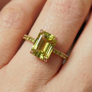May include: A gold ring featuring a large, rectangular, light green gemstone. The band is also gold, with smaller green gemstones set along its surface. The ring is shown on a finger.