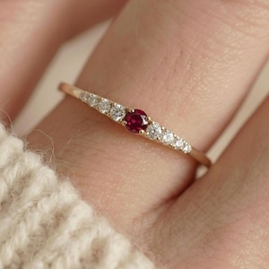 May include: A delicate gold ring featuring a central ruby gemstone, flanked by smaller white gemstones. The ring is set on a finger, showcasing its elegant design and craftsmanship. The ring's slender band and gemstone arrangement create a refined aesthetic.