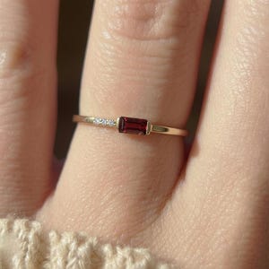 Puede incluir: Un delicado anillo de oro con una piedra preciosa rectangular de color rojo intenso y pequeñas piedras brillantes. La banda del anillo es fina y elegante, mostrando el color vibrante de la piedra central. Una pieza de joyería minimalista.