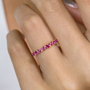 Op de afbeelding: Een gouden ring met een doorlopende rij marquise-geslepen robijnen. De robijnen zijn helderrood en dicht op elkaar gezet. De band van de ring is dun en goudkleurig. De ring wordt getoond op de vinger van een persoon.