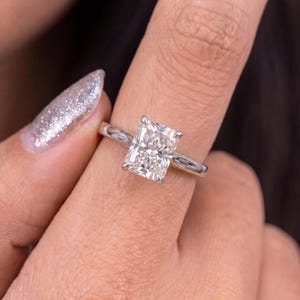 Puede incluir: Un anillo con una piedra preciosa transparente de corte radiante montada en una banda de metal pulido de tono plateado. La piedra central tiene forma rectangular con múltiples facetas que reflejan la luz, sujeta por cuatro garras en una banda lisa.