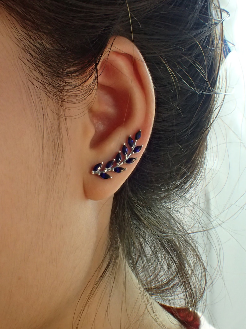 May include: A silver ear cuff with a delicate branch design featuring dark blue gemstones. The cuff is worn on the upper part of the ear.