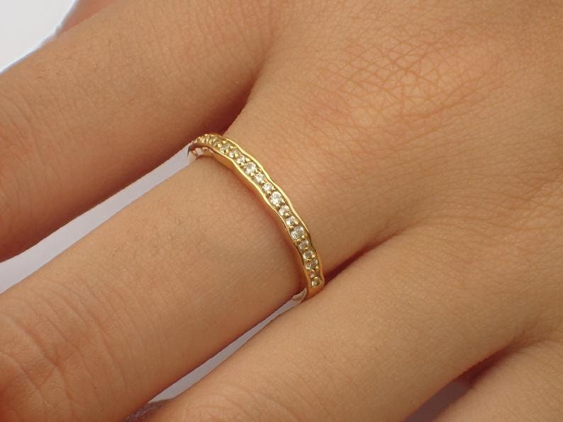 Pu&ograve; includere: Un anello dorato con un design ondulato, incastonato con piccole pietre preziose trasparenti. L'anello &egrave; indossato su un dito, mostrando il suo aspetto delicato ed elegante. Le pietre preziose sono uniformemente distanziate lungo la parte superiore dell'anello.