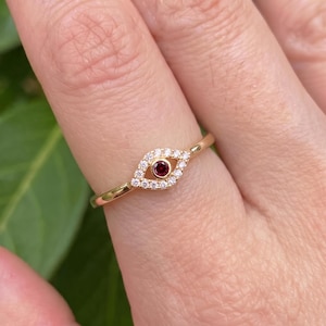 May include: A gold ring featuring an eye-shaped motif centered with a small, round, deep red gemstone. The eye design is outlined with tiny, clear, sparkling stones set into the gold band, creating a delicate and detailed piece of jewelry.