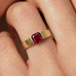 May include: A gold ring featuring a rectangular, deep red gemstone. The ring has a simple, elegant design with a wide band and a bezel setting for the gemstone. The ring is shown on a finger.