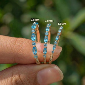 May include: Three gold rings with blue gemstones are displayed. The rings are adorned with oval and round blue gemstones. The text on the image indicates the ring sizes are 2.5mm, 2.0mm, and 1.5mm. The rings are held by a person.