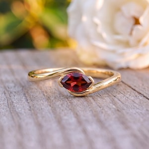 Könnte beinhalten: Ein Goldring mit einem tiefroten Granat im Marquise-Schliff, der in eine Ringschiene im Bypass-Stil gefasst ist. Die polierte Goldschiene schwingt sich um den zentralen Stein, der von kleinen Krappen gehalten wird, und bildet ein Design.