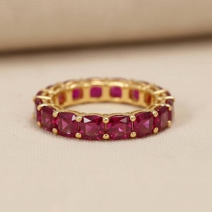 May include: A gold-colored eternity ring featuring square-cut, vibrant pink gemstones. The ring is set with multiple gemstones, creating a continuous band of color. The ring is displayed on a neutral background.