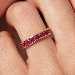 May include: A gold band ring featuring a continuous row of round-cut, vibrant red rubies set in a channel setting. The ring displays a polished metal finish and a series of faceted gemstones that span the width of the band.