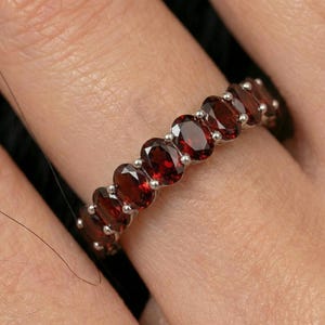Peut inclure: Une bague en argent ornée d'une série de pierres précieuses ovales, rouge foncé. Les pierres sont serties côte à côte, créant une bande continue de couleur. La bague est portée sur un doigt, mettant en valeur le design et la couleur des bijoux.