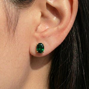 May include: A close-up of a gold stud earring with an oval-shaped, emerald-green gemstone. The earring is set in a four-prong gold setting and is worn on an ear. The background is blurred.
