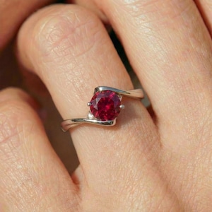 May include: A silver ring with a large, round, deep red gemstone. The band curves around the finger, cradling the stone in a modern, elegant design. The gemstone is the focal point, catching the light and reflecting a rich color.