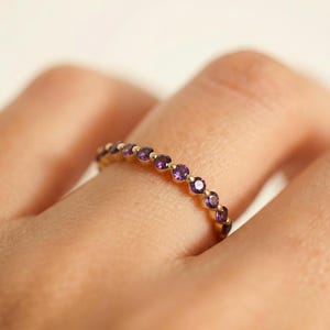May include: A gold ring featuring a series of small, round, purple gemstones. The ring is set in a gold band and is worn on a finger. The gemstones are evenly spaced around the ring.