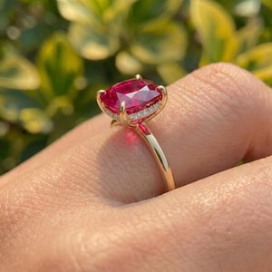 May include: A gold ring featuring a large, cushion-cut, vibrant red gemstone as the centrepiece. The setting includes a halo of small, clear, round-cut stones encircling the base of the main red gem, mounted on a polished, slender gold band.