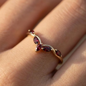 Puede incluir: Un anillo dorado con un diseño curvo que presenta varias piedras preciosas de color rojo intenso con corte marquesa. Las piedras están dispuestas en un delicado patrón en forma de hoja, creando una estética elegante e inspirada en la naturaleza. El anillo se muestra en un dedo.