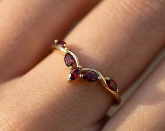 Può includere: Un anello dorato con un design curvo, impreziosito da diverse pietre preziose rosso scuro taglio marquise. Le pietre sono disposte in un delicato motivo a forma di foglia, creando un'estetica elegante e ispirata alla natura. L'anello è mostrato su un dito.