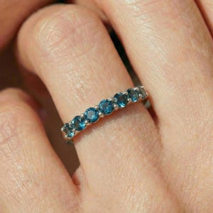 May include: A silver ring with a row of round, vibrant blue gemstones. The gemstones are evenly spaced and set in a band of silver. The ring is shown on a finger, highlighting the jewelry's design and color.