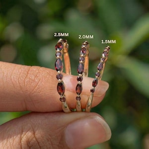 Puede incluir: Se muestran tres anillos dorados con piedras preciosas rojas. Los anillos están etiquetados como 2,5 mm, 2,0 mm y 1,5 mm. Cada anillo presenta una serie de piedras preciosas rojas ovaladas y redondas engastadas a lo largo de la banda. Los anillos se sostienen sobre un fondo verde borroso.