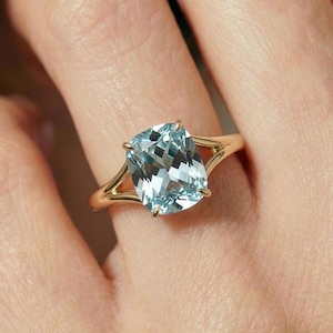 Puede incluir: Un anillo de oro con una gran gema azul claro con talla de cojín. La banda del anillo se divide en un diseño de doble banda a cada lado de la gema, creando un aspecto único y elegante. La gema se sujeta con cuatro garras.