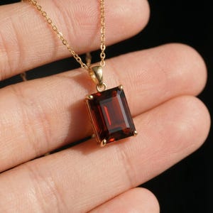 May include: A gold necklace with a rectangular, deep red gemstone pendant. The pendant is set in a gold frame and hangs from a delicate gold chain. The gemstone has a rich, polished appearance.