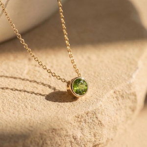 May include: A gold-colored necklace with a round, light green gemstone pendant. The pendant is set in a gold bezel and hangs from a delicate chain. The necklace is displayed on a textured, light brown surface.