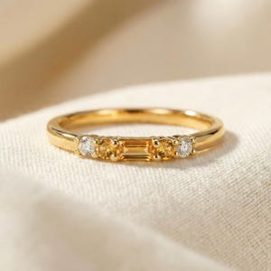 Könnte beinhalten: Ein goldener Ring mit einer Reihe von Edelsteinen. Der Ring hat einen zentralen rechteckigen gelben Edelstein, der von kleineren runden gelben Edelsteinen und klaren, runden Edelsteinen flankiert wird. Der Ring liegt auf einem weißen Stoff.