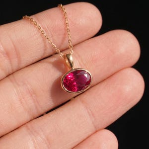 May include: A gold-toned necklace featuring a faceted, oval-shaped, deep pink gemstone pendant set in a smooth gold-toned bezel. The pendant hangs from a delicate, matching gold-toned chain, showcasing a vibrant, reflective surface.
